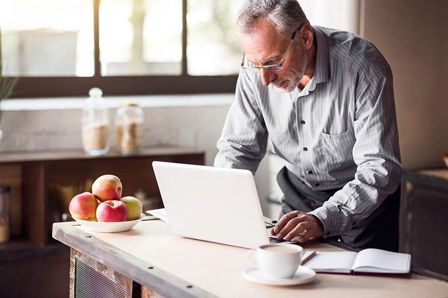 Blog - Older Man Standing in Kitchen While Looking and Typing on a Laptop with Apples Sitting on the Counter Next to Him